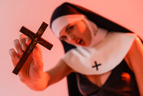 angry nun holding crucifix while screaming on blurred foreground isolated on pink
