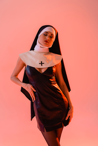 sexy nun in black leather dress posing with hand on hip and closed eyes isolated on pink