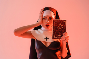 shocked nun in sexy dress touching head while holding jewish bible isolated on pink