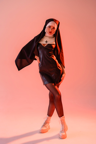 young nun in black leather dress posing with hand on hip on pink background