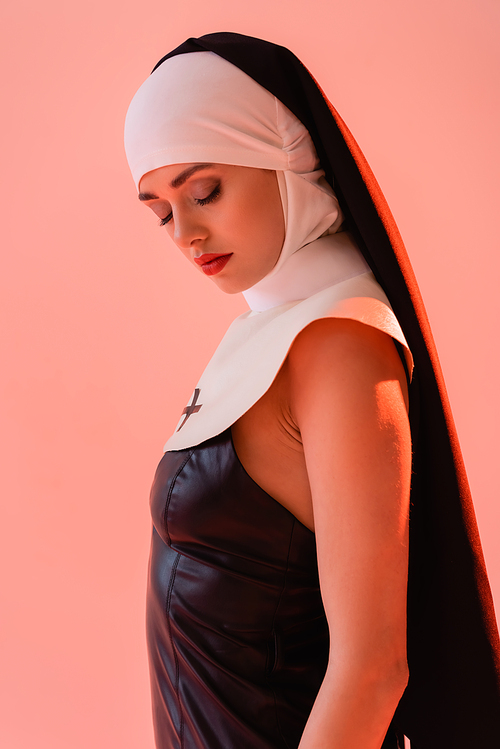 sensual nun in leather dress standing with closed eyes isolated on pink