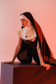 young nun in sexy dress looking away while sitting on black cube isolated on pink