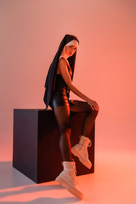 sexy nun in leather dress and boots posing on black cube on pink background