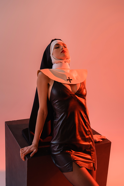 sensual nun in sexy dress posing with closed eyes near black cube on pink background