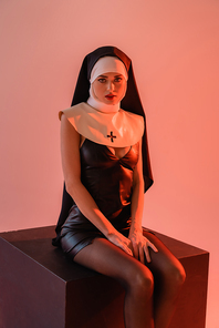 sexy nun in leather dress  while sitting on black cube isolated on pink