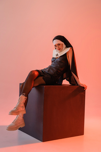 young nun in sexy dress and white boots sitting on black cube on pink background