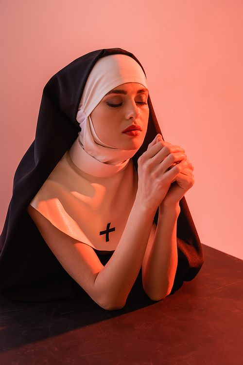 young nun praying with closed eyes on black surface isolated on pink