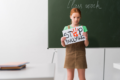 selective focus of sad kid holding placard with stop bullying lettering in classroom