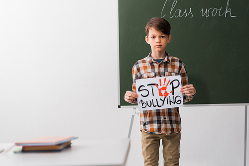 selective focus of schoolboy holding placard with stop bullying lettering in classroom