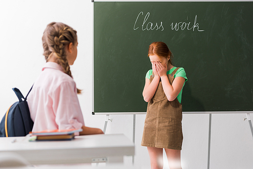 selective focus of schoolkid covering face while crying near classmate, bullying concept