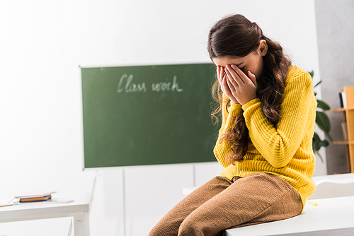 upset and bullied schoolgirl covering face while crying in classroom