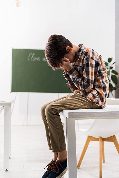 bullied schoolchild covering face while crying in classroom