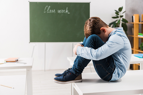 bullied schoolchild covering face while crying and sitting on table