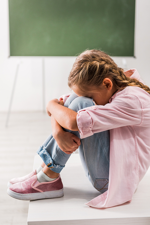 upset and bullied schoolgirl covering face while sitting on desk in classroom