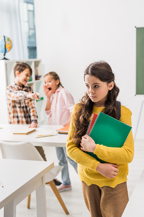 selective focus of bullied schoolgirl holding notebooks near cruel classmates