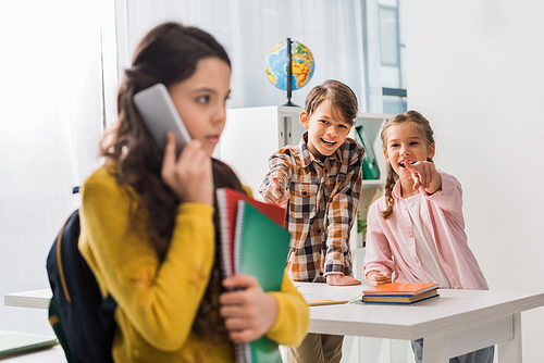 selective focus of cruel classmate pointing with finger at upset schoolgirl talking on smartphone, bullying concept