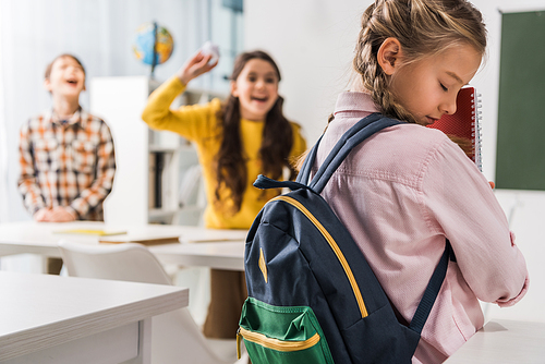 selective focus of bullied schoolgirl with backpack holding notebooks near cruel schoolkids