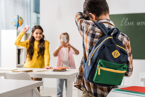 back view of bullied kid standing near cruel classmates with smartphone, cyberbullying concept