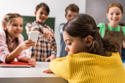 selective focus of bullied schoolgirl sitting near schoolkid with smartphone and cruel  classmates, cyberbullying concept