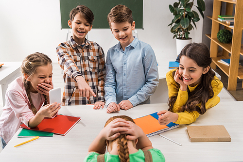 selective focus of schoolkid with smartphone and cruel classmates laughing near bullied kid, cyberbullying concept