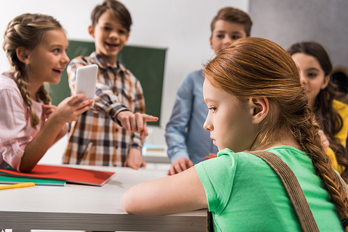 selective focus of depressed schoolgirl sitting near schoolkid with smartphone and cruel classmates, cyberbullying concept