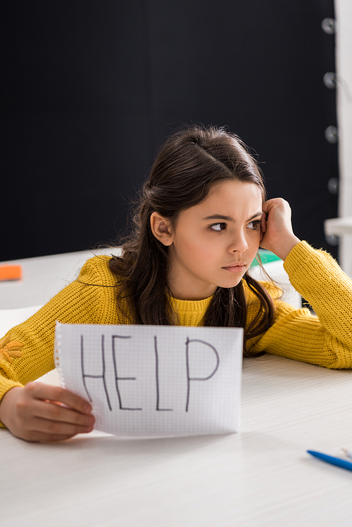 upset schoolgirl holding paper with help lettering and looking away, bullying concept