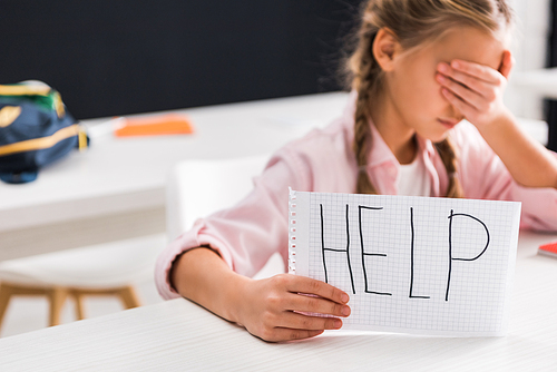 selective focus of paper with help lettering in hand of upset schoolgirl covering eyes, bullying concept