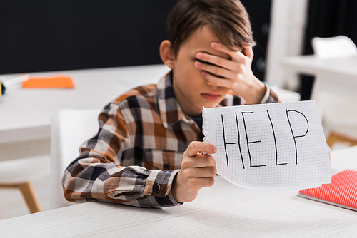 selective focus of upset schoolboy holding paper with help lettering and covering eyes, bullying concept