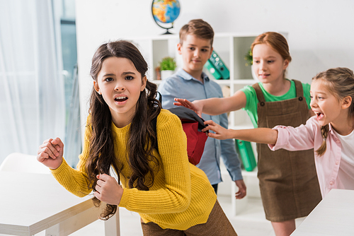 selective focus of cruel schoolkids touching backpack of bullied schoolgirl