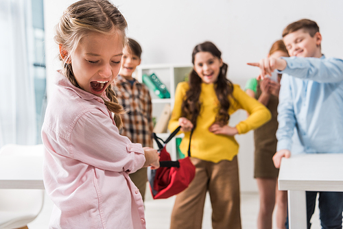 selective focus of bullied schoolgirl screaming near cruel cruel pupils holding backpack and pointing with fingers