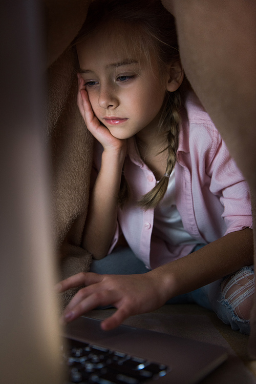 selective focus of frustrated kid covered in blanket using laptop, cyberbullying concept