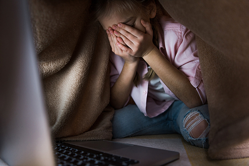 selective focus of frustrated kid covered in blanket covering face while crying near laptop, cyberbullying concept