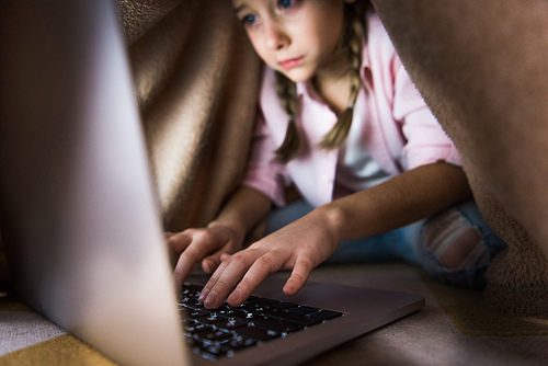 selective focus of frustrated kid covered in blanket typing on laptop keyboard, cyberbullying concept