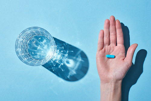 cropped view of woman holding blue pill on palm near glass with water on blue background