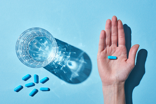 partial view of woman holding blue pill on palm near glass with water on blue background