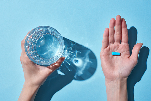 cropped view of woman holding blue pill on palm and glass with water on blue background