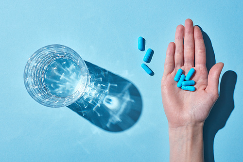 cropped view of woman holding blue pills on palm near glass with water on blue background