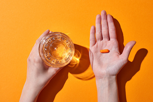 cropped view of woman holding pill on palm near glass with water on orange background