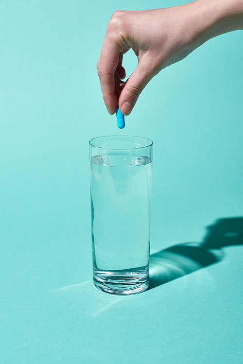 cropped view of woman putting blue pill in transparent glass with fresh water on turquoise background