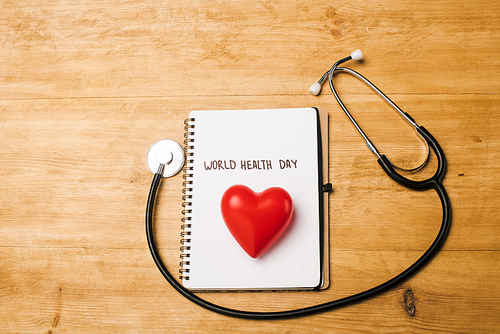 Top view of stethoscope, decorative heart on notebook with world health day lettering on wooden background