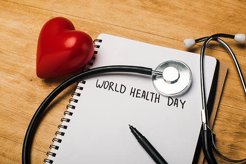 Stethoscope, decorative heart, pen on notebook with world health day lettering on wooden background