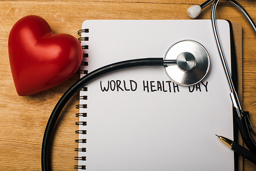 Top view of decorative heart, stethoscope, pen on notebook with world health day lettering on wooden background