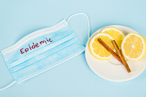 top view of saucer with cinnamon sticks and lemons near medical mask with epidemic lettering on blue