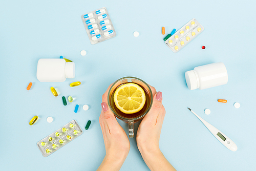 top view of woman holding cup of tea with sliced lemon near pills and digital thermometer on blue