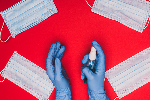 Top view of doctor in latex gloves holding hand sanitizer near medical masks on red background