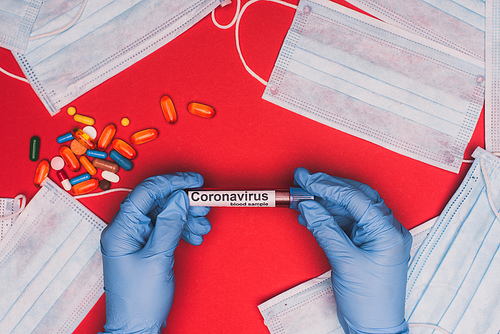 Top view of doctor holding test tube with blood sample and coronavirus lettering near pills and medical masks on red background