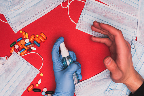 Cropped view of man holding bottle of hand sanitizer near pills and medical masks on red surface