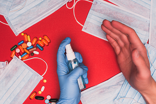 Top view of man in latex glove using hand sanitizer near medical masks with pills on red background