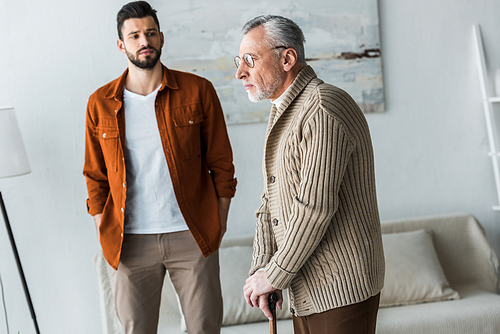 handsome bearded son standing with hands in pockets and looking at sad senior father in glasses