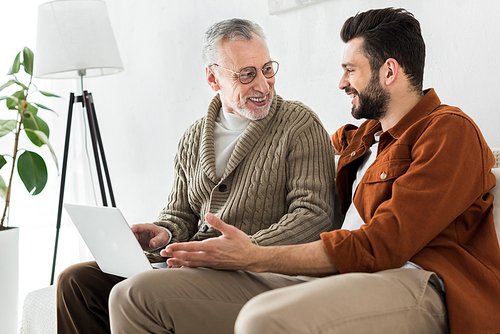 happy bearded man gesturing while sitting with senior father and looking at laptop